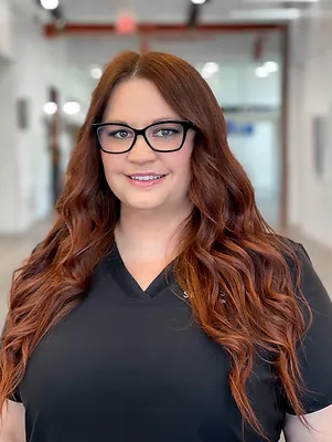 Portrait of Michelle Crandall, a smiling woman with long, wavy red hair, wearing black glasses and a black top, standing in a modern office setting.